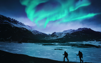 Hikers On A Frozen Lake, Mountains And Northern Lights In The Background