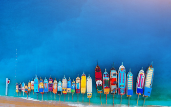 Row of colourful boats along a sandy blue beach taken in an aerial shot